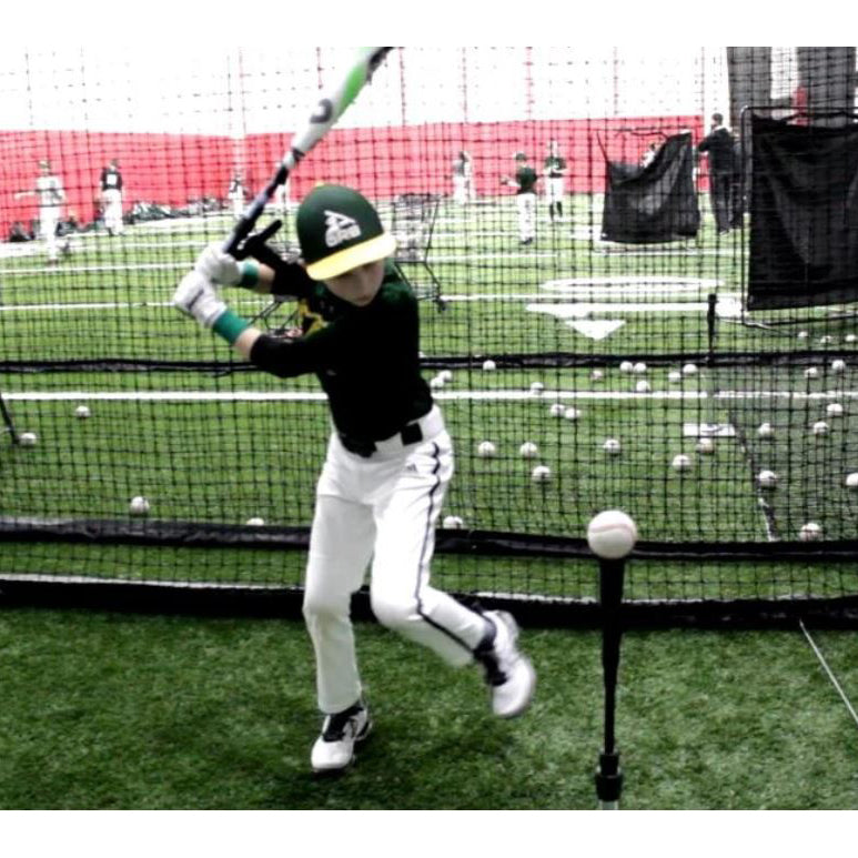 A young baseball player in a green uniform practices hitting a ball on a tee using the SWINGRAIL Baseball & Softball Swing Trainer by SWINGRAIL in an indoor facility, with scattered baseballs on the artificial turf behind him.