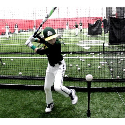 A young baseball player in a green uniform practices hitting a ball on a tee using the SWINGRAIL Baseball & Softball Swing Trainer by SWINGRAIL in an indoor facility, with scattered baseballs on the artificial turf behind him.