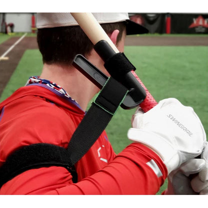 A baseball player in a red shirt and white gloves uses the SWINGRAIL Baseball & Softball Swing Trainer by SWINGRAIL, strapped to his arm and bat, to improve swing mechanics and bat speed on a green indoor field.