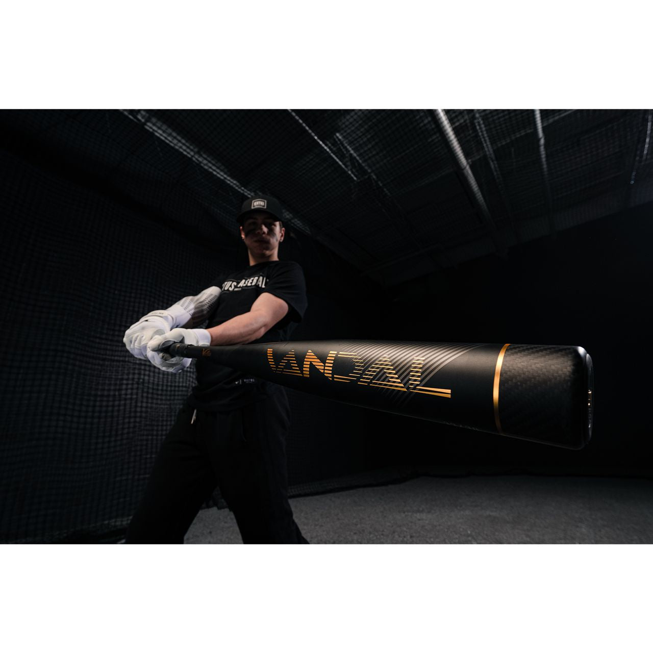 A baseball player in a black cap, black shirt, and white gloves holds a Victus 2022 Vandal 2 (-8) USSSA Baseball Bat (VSBV2X8) toward the camera in a dark indoor batting cage.