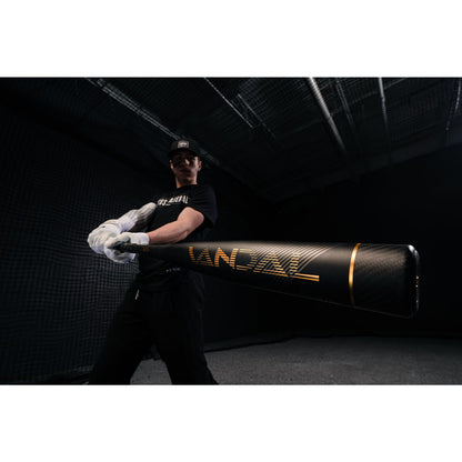A baseball player in a black cap, black shirt, and white gloves holds a Victus 2022 Vandal 2 (-8) USSSA Baseball Bat (VSBV2X8) toward the camera in a dark indoor batting cage.