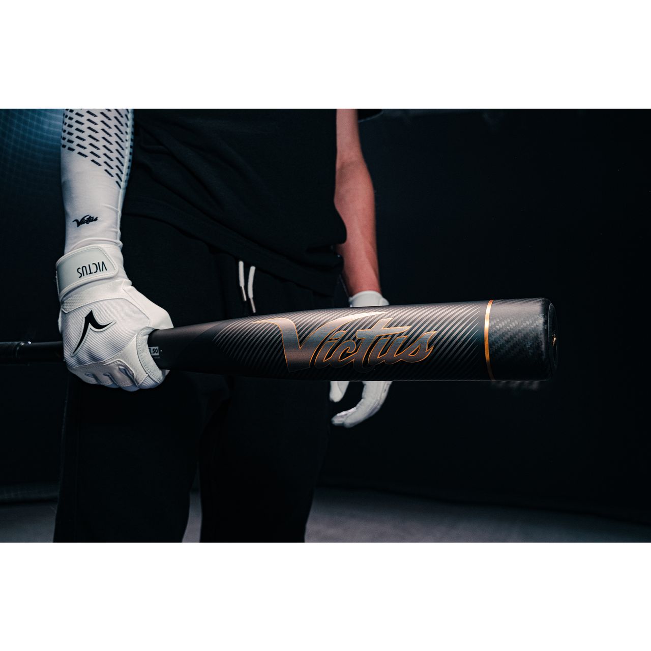 Wearing white batting gloves, a person holds a Victus Vandal 2 (-8) USSSA Baseball Bat (2022, black/gold) with one hand in a dark setting; only their upper body is partially seen.