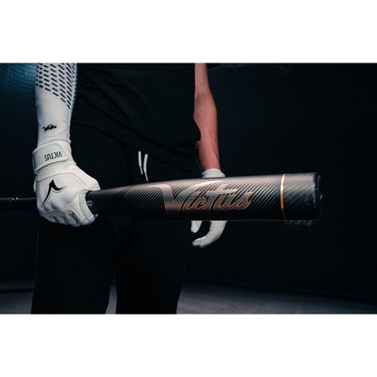 Wearing white batting gloves, a person holds a Victus Vandal 2 (-8) USSSA Baseball Bat (2022, black/gold) with one hand in a dark setting; only their upper body is partially seen.