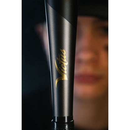 A close-up of a black 2022 Victus Vandal 2 (-8) 2 3/4" USSSA Baseball Bat by Victus, featuring gold lettering, held vertically in front of a person with blurred facial features and black eye paint.
