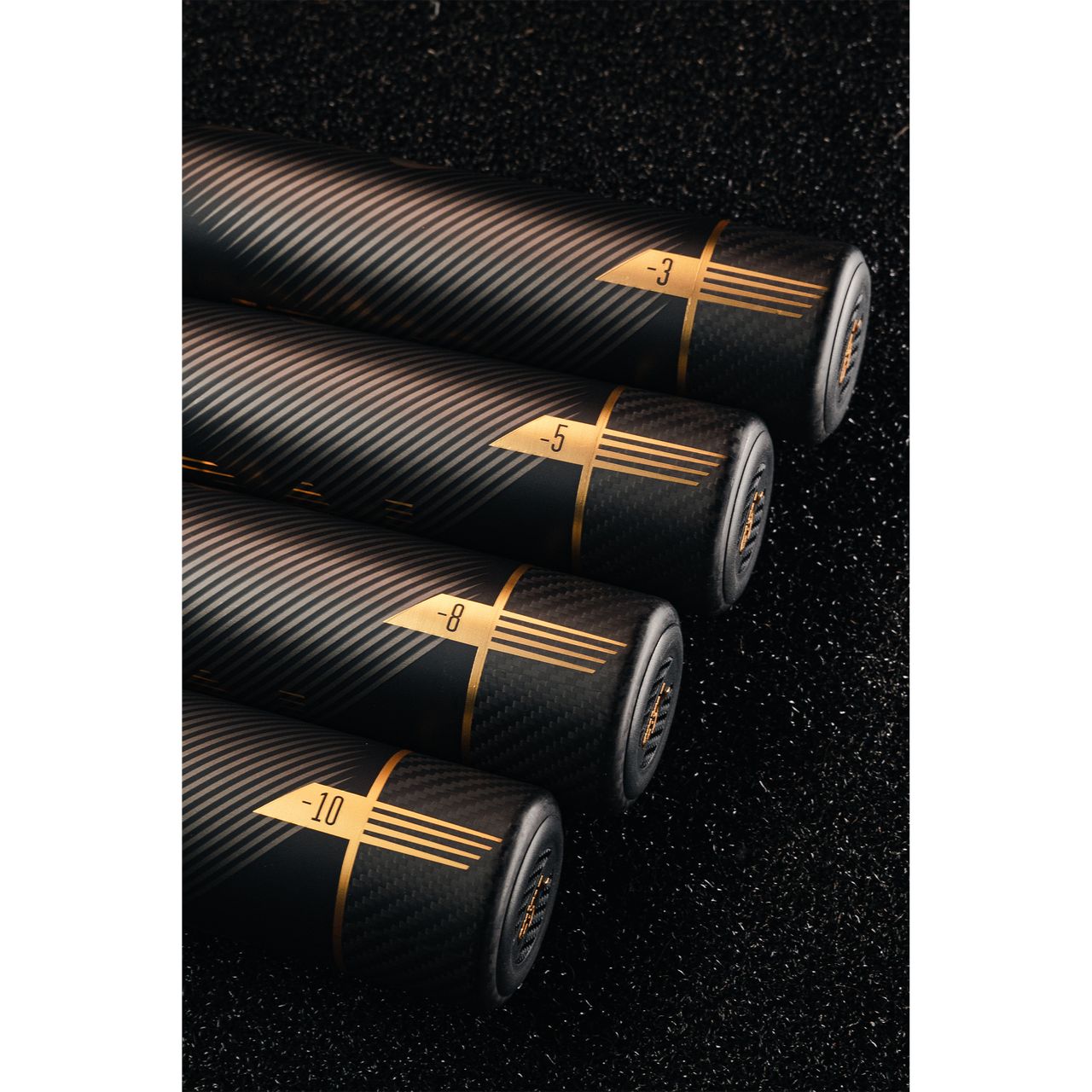 Four black cylindrical objects with gold accents and numbers (-3, -5, -8, -10) are arranged in a row on a textured black surface, featuring a sleek carbon fiber design like the 2022 Victus Vandal 2 (-8) USSSA Baseball Bat by Victus.