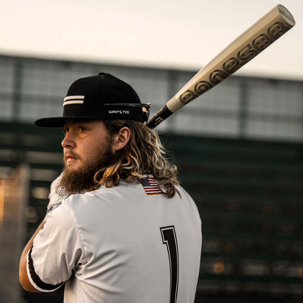 A baseball player with long hair and a beard, wearing a white jersey numbered 1 and cap, holds a Warstic 2024 Bonesaber Hybrid (-3) BBCOR Baseball Bat (MBBSRHB23WH3 DEMO) over his shoulder. An American flag patch is on his back; empty bleachers are behind him.