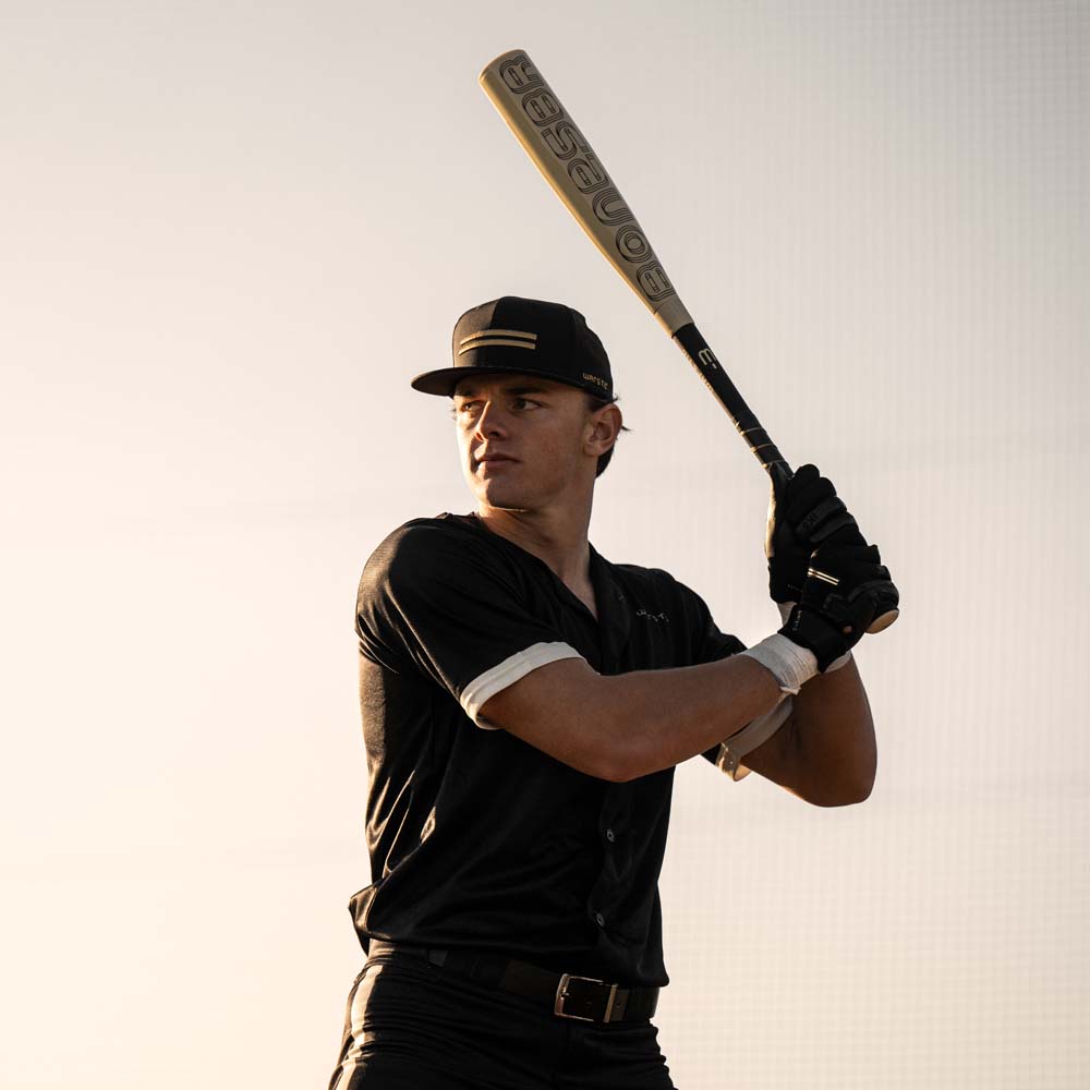 A baseball player stands ready to bat, gripping a 2024 Warstic Bonesaber Hybrid (-3) BBCOR Baseball Bat: MBBSRHB23WH3 (DEMO) over his shoulder. He's wearing a black uniform, cap, and gloves, looking focused against a light background.