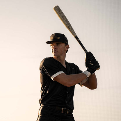 A baseball player stands ready to bat, gripping a 2024 Warstic Bonesaber Hybrid (-3) BBCOR Baseball Bat: MBBSRHB23WH3 (DEMO) over his shoulder. He's wearing a black uniform, cap, and gloves, looking focused against a light background.