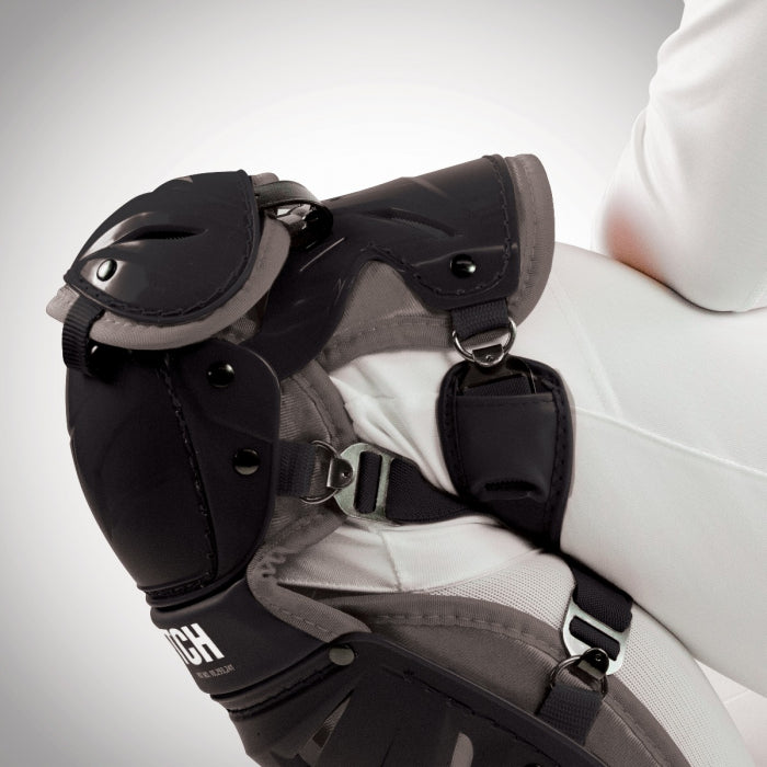 Close-up of a person in equestrian gear, highlighting All Star AFx Fastpitch Catcher's Leg Guards: LGW-AFX worn over white riding pants, featuring detailed straps and buckles for added protection, similar to traditional catcher leg guards.