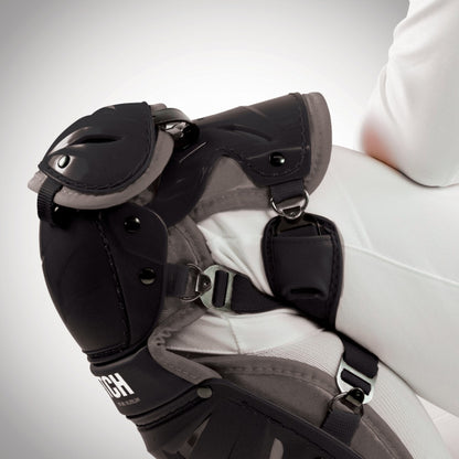 Close-up of a person in equestrian gear, highlighting All Star AFx Fastpitch Catcher's Leg Guards: LGW-AFX worn over white riding pants, featuring detailed straps and buckles for added protection, similar to traditional catcher leg guards.