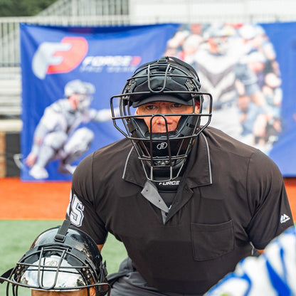 An umpire in a Force3 Pro Gear Traditional Defender Mask: BD31 crouches behind the catcher on the field, with blurred promotional banners and stands in the background.