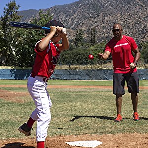 A coach tosses PowerNet 3.2" Weighted Hitting and Batting Training Balls (6 Pack: 1004L) to a young player at bat, both in red shirts and white pants, focusing on building batting power and arm strength under sunny skies.