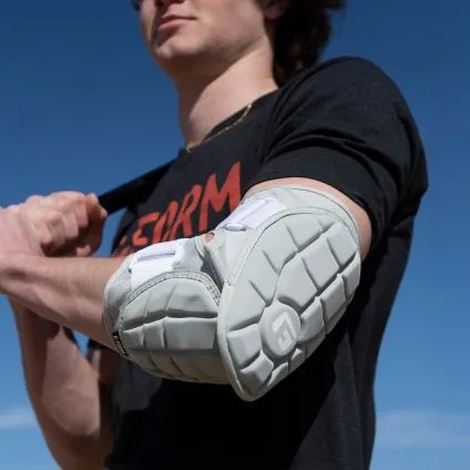 A person in a black shirt with red text uses the G-Form Elite 2 Batter's Elbow Guard (EP1410) by G-Form as an improvised pad, gripping its strap over their shoulder against a clear blue sky.