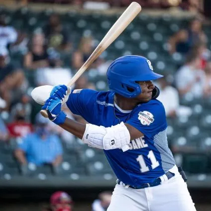 A baseball player in a blue uniform and helmet, wearing number 11, grips his bat and prepares to hit while protected by the G-Form Elite 2 Batter's Elbow Guard (EP1410) from G-Form. Spectators watch from the blurred background.