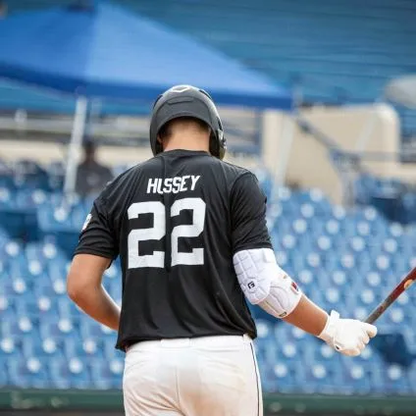 A baseball player in a black jersey labeled Hissey (#22) stands with a bat, wearing the G-Form Elite 2 Batter's Elbow Guard (EP1410) by G-Form, in a stadium with empty blue seats in the background.