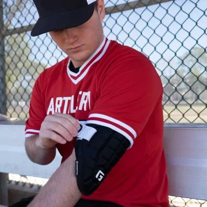A baseball player in a red jersey and black cap sits on a bench by a chain-link fence, adjusting the G-Form Elite Speed Batter's Elbow Guard: EP15 on his right arm.