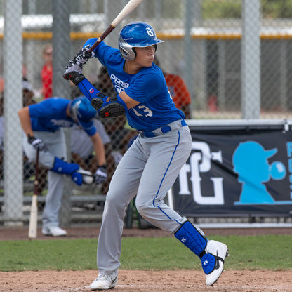 A baseball player in a blue uniform, helmet, and G-Form Batter's Leg Guard (LG0102) stands at bat, focused on the pitcher. A teammate appears in the background with a fence and banner behind them.