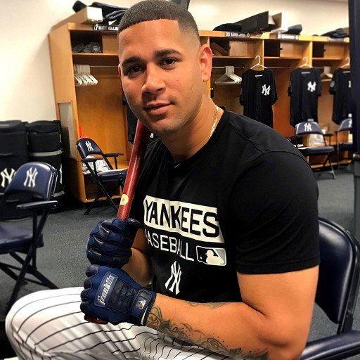 A Yankees player sits in a locker room holding a bat and wearing Franklin CFX Pro Full Color Chrome Adult Batting Gloves by Franklin, along with striped pants. Lockers, chairs, and jerseys are visible in the background.