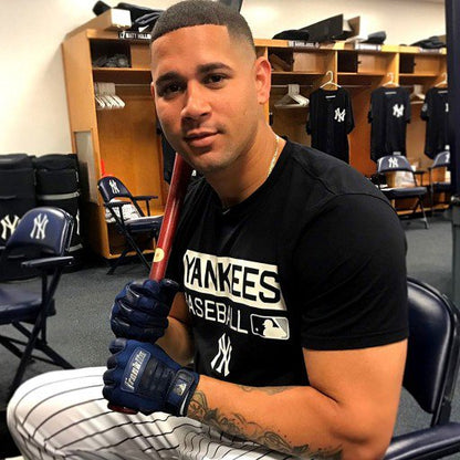 A Yankees player sits in a locker room holding a bat and wearing Franklin CFX Pro Full Color Chrome Adult Batting Gloves by Franklin, along with striped pants. Lockers, chairs, and jerseys are visible in the background.