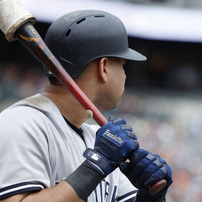 A baseball player in a black helmet and gray uniform holds a bat on his shoulder, wearing Franklin CFX Pro Full Color Chrome Adult Batting Gloves for superior grip. The stadium crowd appears blurred in the background.