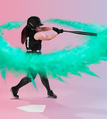 A softball player in a black uniform and Rip It Vision Pro Home Fastpitch Softball Batting Helmet with Mask swings, sending vibrant green powder around them over home plate, with a background blending pink to purple.