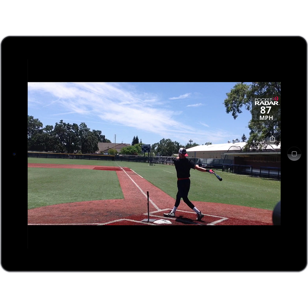 A baseball player swings a bat at home plate, with the Pocket Radar Smart Coach Radar: SR1100 by Pocket Radar displaying an 87 MPH speed on a tablet. The sky is clear, and trees and buildings fill the background.