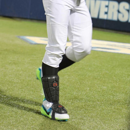 A baseball player wearing white pants, black socks, green cleats, and a G-Form Batter's Leg Guard (LG0102) with SmartFlex padding stands on a grass field.