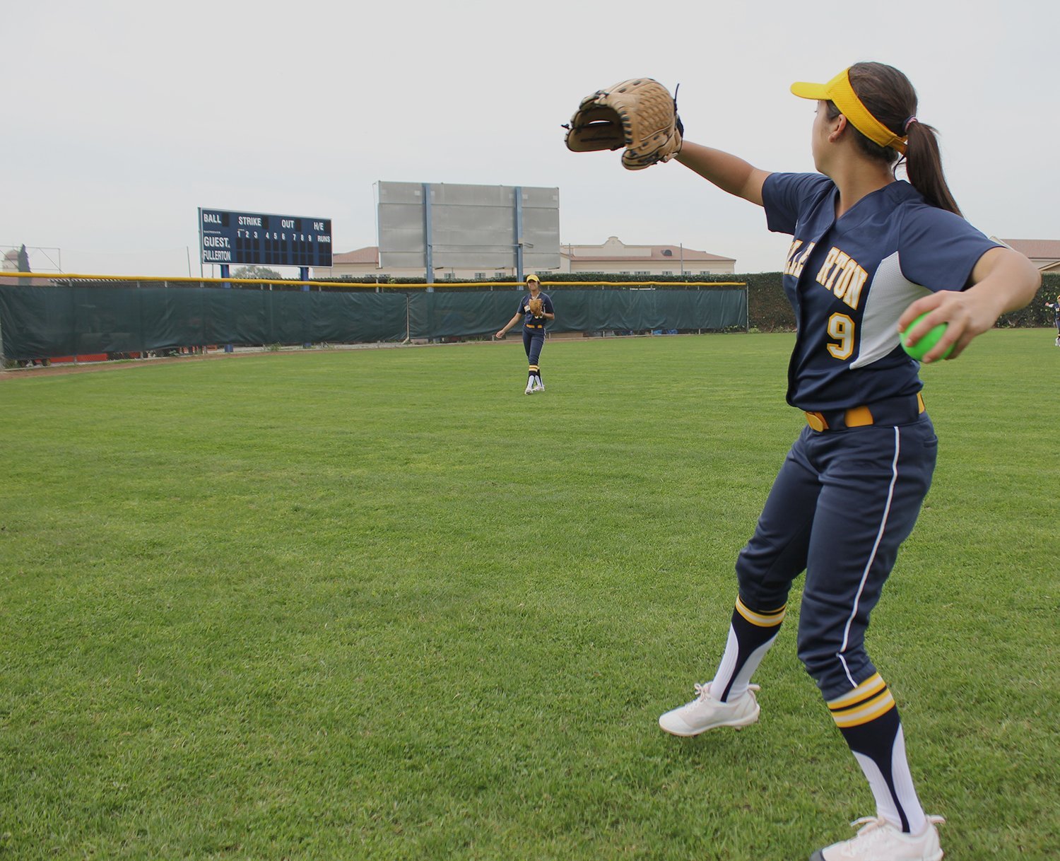 Two female softball players in navy uniforms practice throwing PowerNet 2.8" Weighted Hitting and Batting Progressive Training Balls (9 Pack) on a grassy field; one is mid-throw with a glove up, scoreboard in the background.