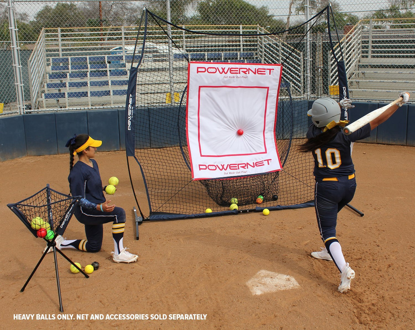 Two softball players use PowerNet 2.8" Weighted Hitting and Batting Progressive Training Balls (9 Pack) in practice; one kneels by a basket of yellow balls, while the other swings at a ball toward a net with empty bleachers in the background.
