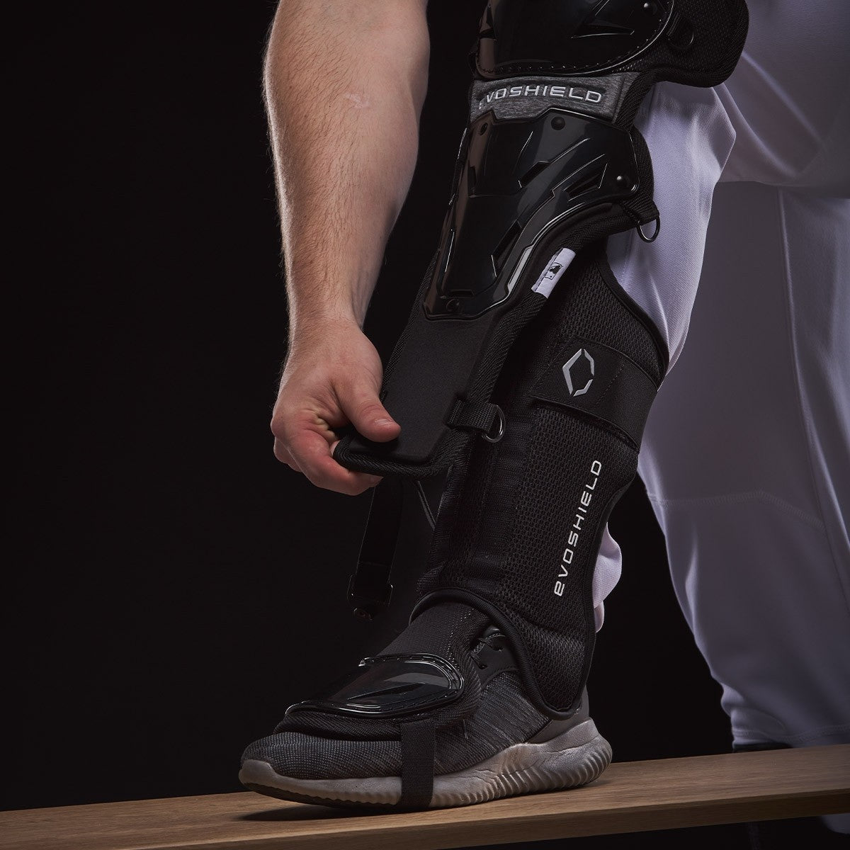 A person in white pants fastens EvoShield Pro-SRZ Gel-to-Shell™ Catcher's Lower Leg Guards (WB570800 / WB571030) over their shin and foot, standing on a wooden bench against a dark background.