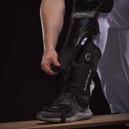 A person in white pants fastens EvoShield Pro-SRZ Gel-to-Shell™ Catcher's Lower Leg Guards (WB570800 / WB571030) over their shin and foot, standing on a wooden bench against a dark background.