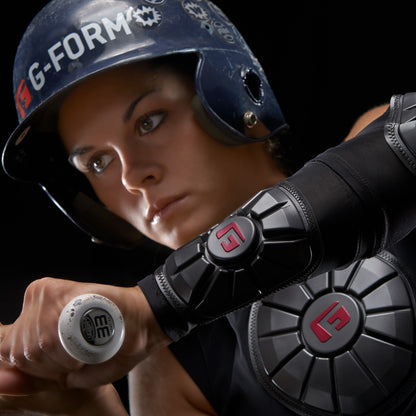 A focused athlete wearing a blue helmet and black gear with red G-Form logos grips a bat, highlighting the advanced impact protection of the G-Form Pro Wrist Guard (WG0102) with visible SmartFlex padding on the arm and chest.