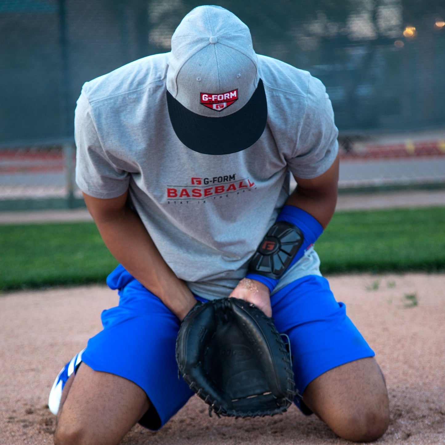 A baseball player in a gray shirt and blue shorts kneels on the field holding his groin. He wears a glove, a G-Form Pro Wrist Guard: WG0102 by G-Form for impact protection, and a protective arm guard. A fence and grass are in the background.
