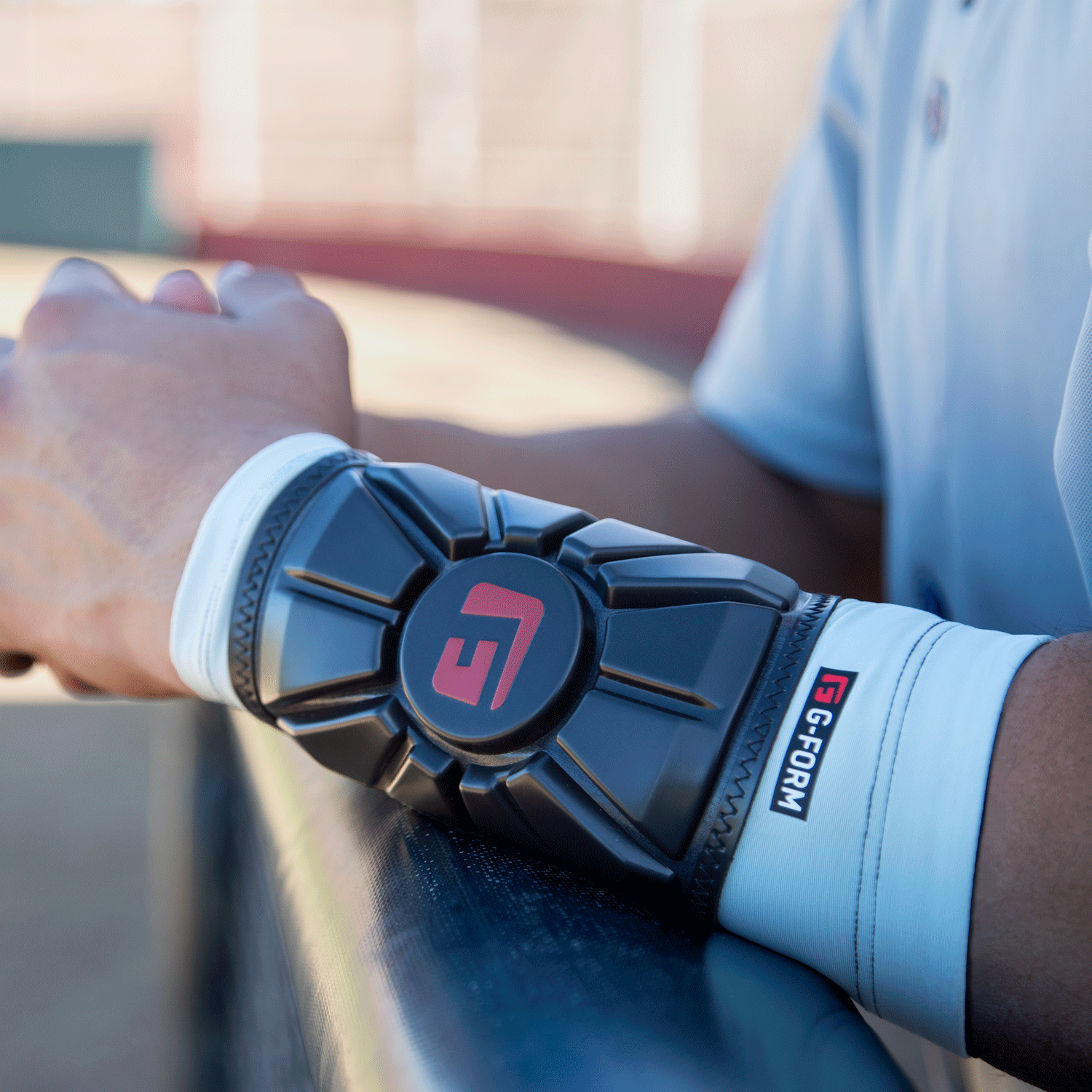 Wearing a white shirt, a person leans on a rail and displays the G-Form Pro Wrist Guard WG0102 with SmartFlex protection, as a blurred sports field fills the background.